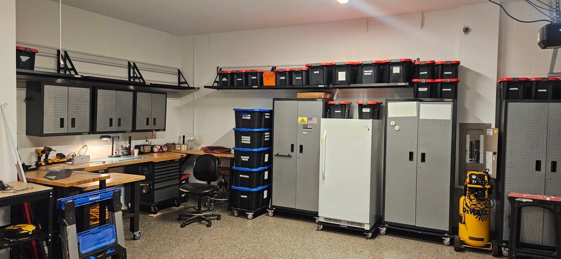Garage with wall-mounted shelves, storage bins and cabinets above a workbench.